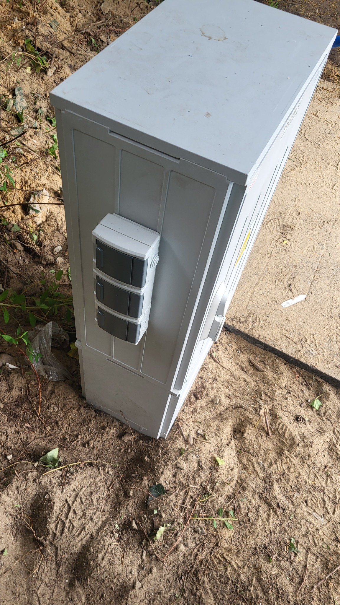 Szara szafka elektryczna z trzema zabezpieczeniami zamontowana na zewnątrz, stojąca na ziemi z piaskiem i roślinnością.