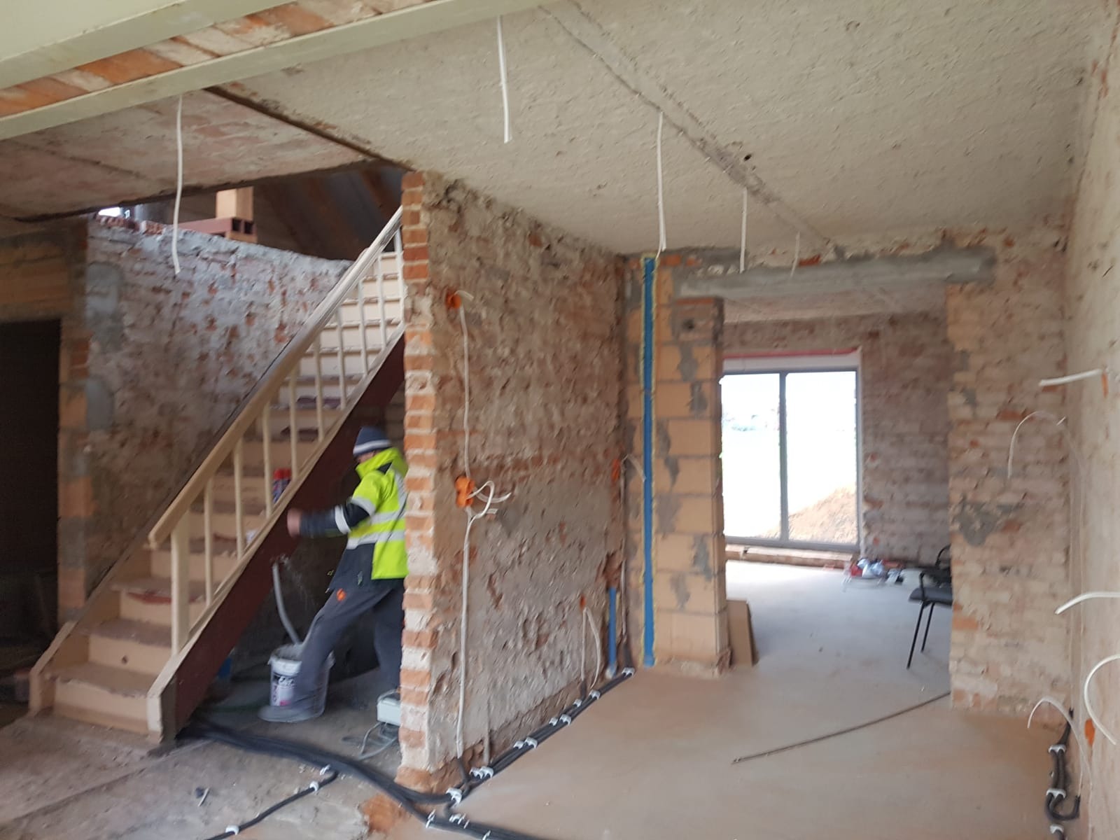 Wnętrze w trakcie remontu: instalacja przewodów elektrycznych przy podłodze, widoczne puszki elektryczne w ścianach z cegły, pracownik w odblaskowej kamizelce przy schodach.