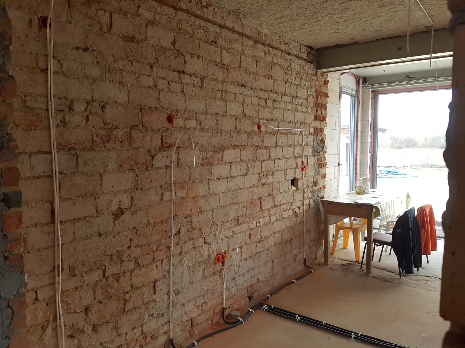 Surowa ceglana ściana z poprowadzonymi przewodami elektrycznymi w peszlach i pomarańczowych puszkach instalacyjnych, widoczne przygotowania do prac wykończeniowych wnętrza i instalacji elektrycznej.