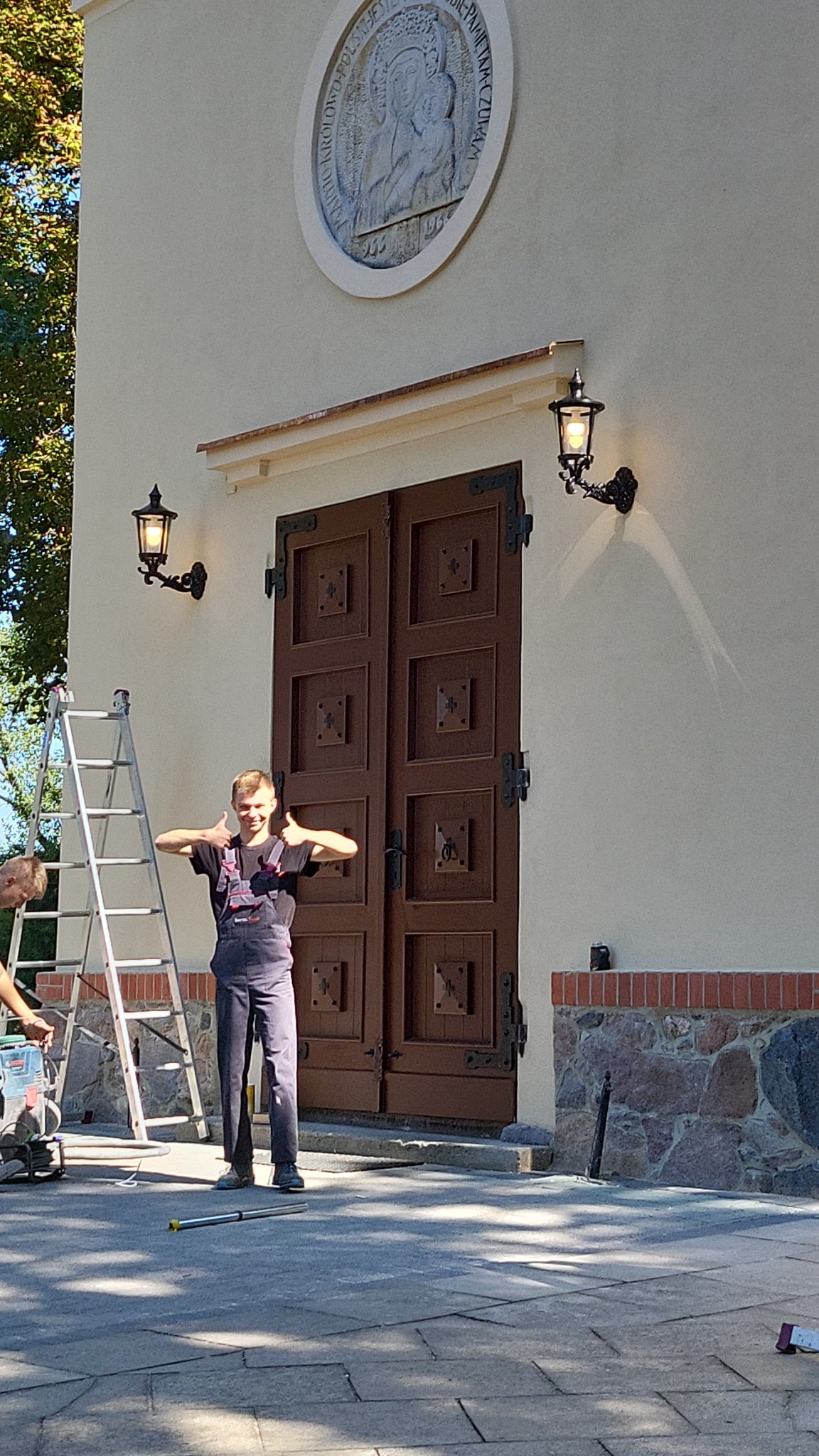 Młody elektryk w kombinezonie, z uśmiechem prezentuje zamontowane lampy zewnętrzne przy dużych, drewnianych drzwiach kościoła, z widoczną drabiną i narzędziami w tle.