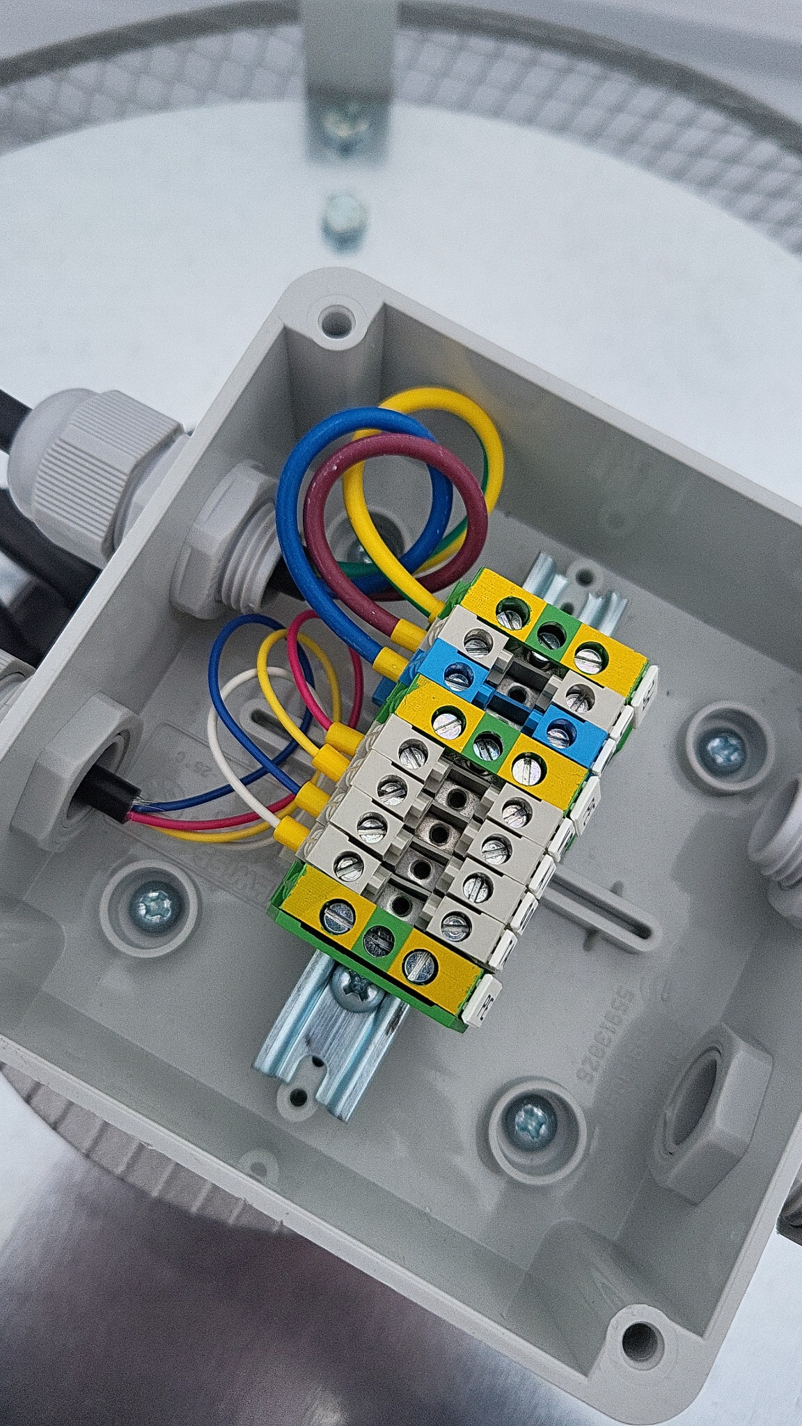 Wnętrze szarej puszki instalacyjnej z listwą zaciskową, widoczne kolorowe przewody podłączone do listwy i wchodzące przez dławiki.