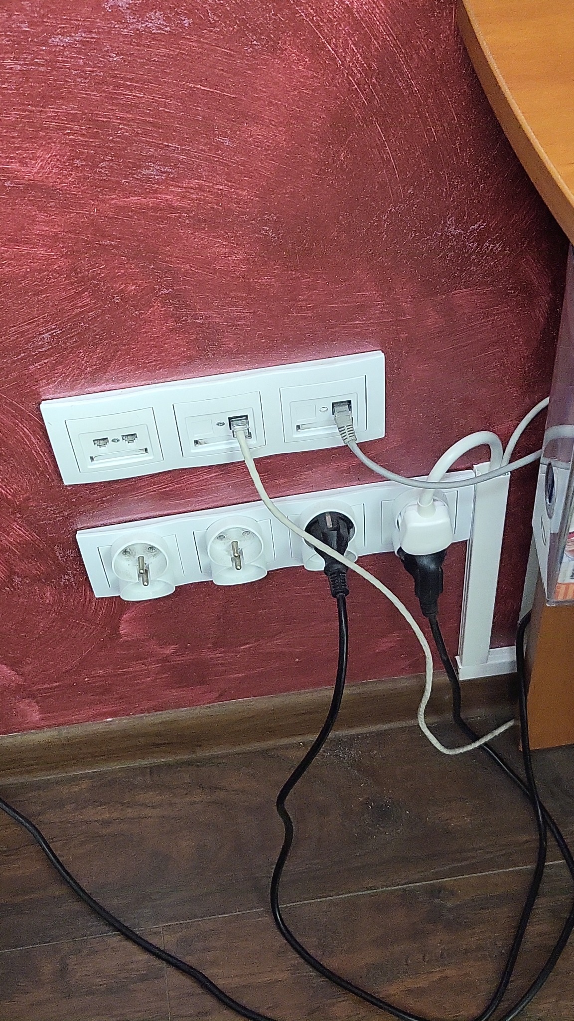 Gniazda elektryczne i internetowe w białych ramkach, zamontowane na bordowej ścianie z widoczną fakturą, z wpiętymi wtyczkami i kablami.