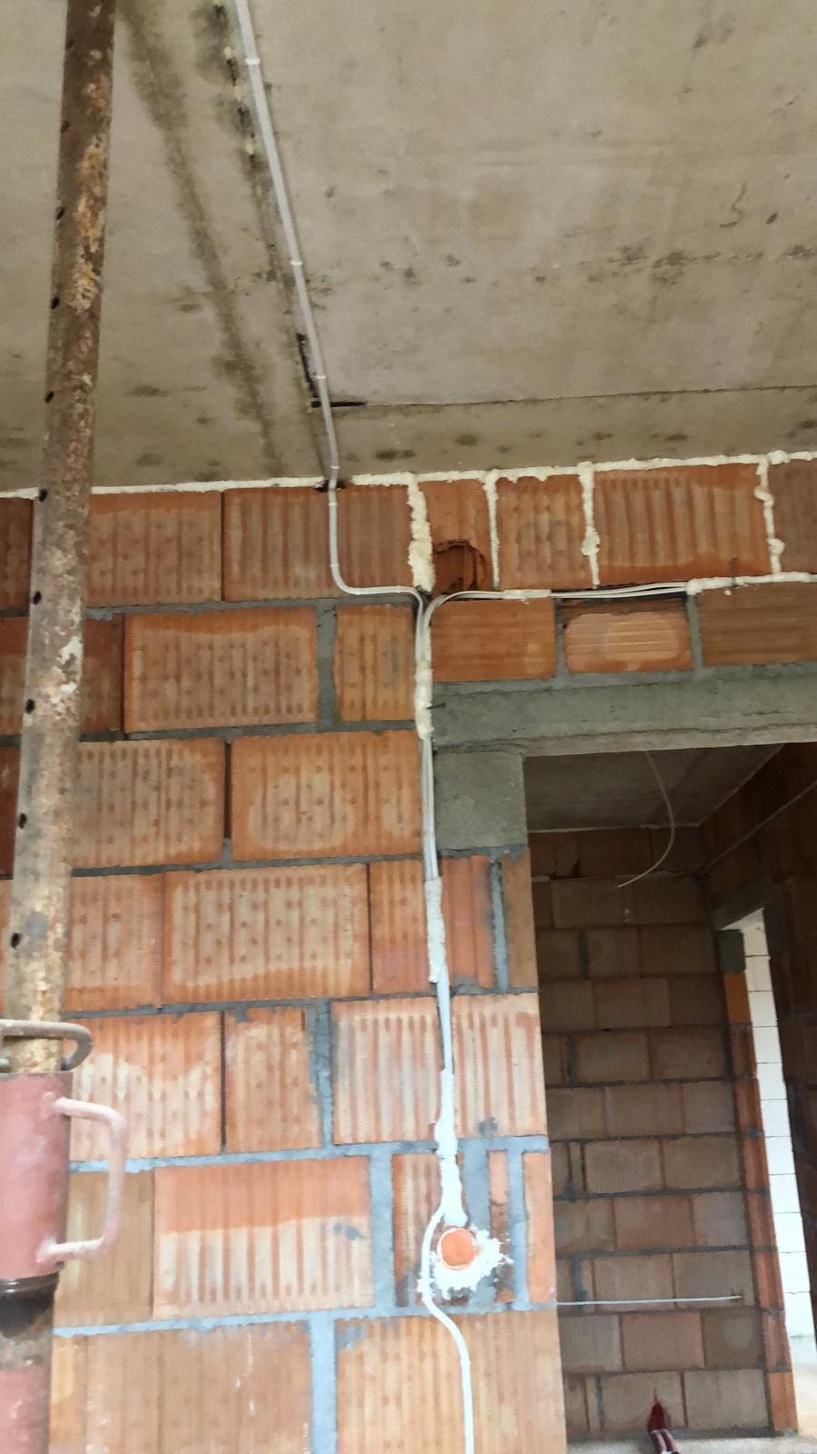 Surowy stan: Białe kable elektryczne poprowadzone po ścianie z czerwonej cegły dziurawki i pod betonowym sufitem w trakcie budowy, widoczna puszka elektryczna i fragment stalowego stempla budowlanego.