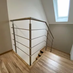 Nowoczesne schody z drewnianymi stopniami i metalową balustradą z poziomymi prętami, oświetlone światłem wpadającym przez okno dachowe.