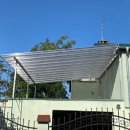 Zadaszenie tarasu z przezroczystych paneli zamontowane przy jasnym budynku z zielonymi elementami, widoczne zza ozdobnego metalowego ogrodzenia.