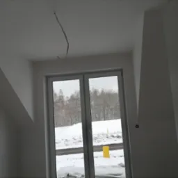 Poddasze po malowaniu na biało z widocznym kablem wystającym z sufitu i oknem z widokiem na zaśnieżony krajobraz.