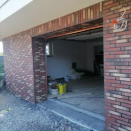 W trakcie prac: Nowa elewacja garażu wykończona czerwoną cegłą klinkierową, widoczne narzędzia i materiały budowlane wewnątrz garażu, teren wokół garażu jeszcze niezagospodarowany.