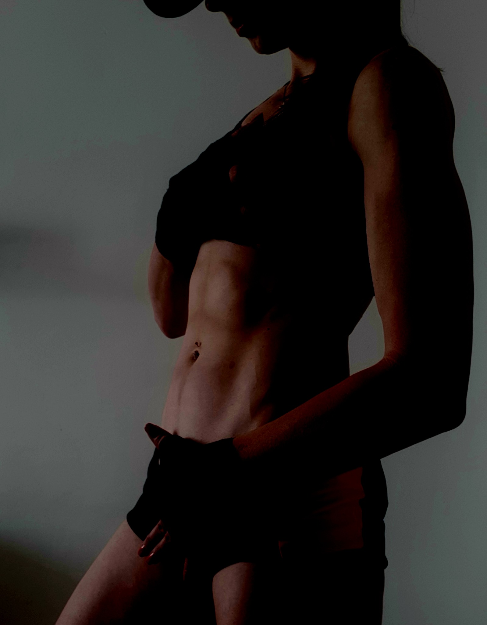 Silna, wyrzeźbiona sylwetka kobiety w sportowym stroju, z widocznymi mięśniami brzucha, pozująca na tle ściany w stonowanym oświetleniu.