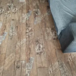 Panele podłogowe z efektem postarzanego drewna i nadrukami w stylu vintage, częściowo zasłonięte przez szarą sofę.