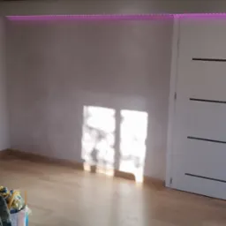 Pok&oacute;j po odświeżeniu z nową podłogą, pomalowanymi ścianami w odcieniach szarości, białą stolarką drzwiową z poziomymi wstawkami i listwą LED RGB pod sufitem, widoczne narzędzia i drabina malarska.