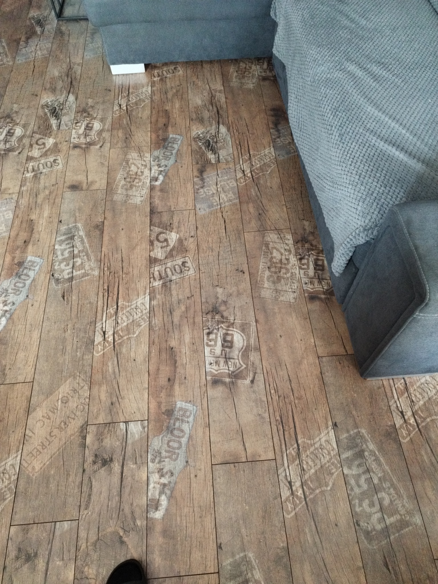 Panele podłogowe z efektem postarzanego drewna i nadrukami w stylu vintage, częściowo zasłonięte przez szarą sofę.