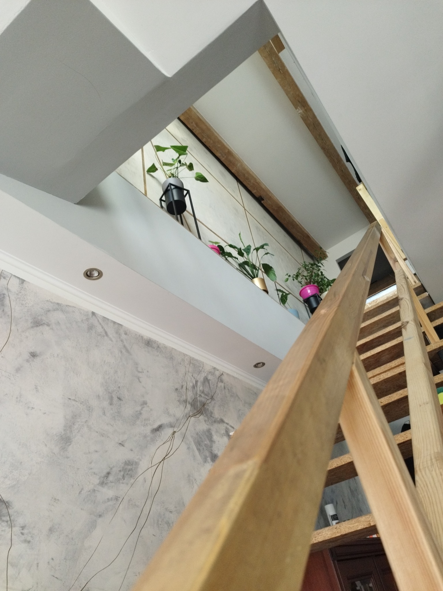 Widok z dołu na drewniane schody z surowego drewna, prowadzące do otworu w suficie, przez który widać ścianę z dekoracyjną szarą fakturą i rośliny doniczkowe na półce.