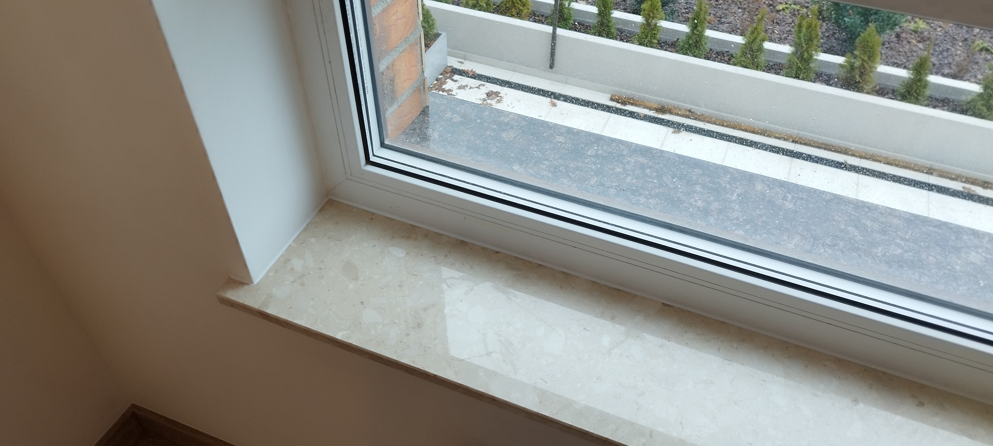 Marmurowy parapet wewnętrzny pod oknem z widokiem na balkon z roślinami iglastymi.