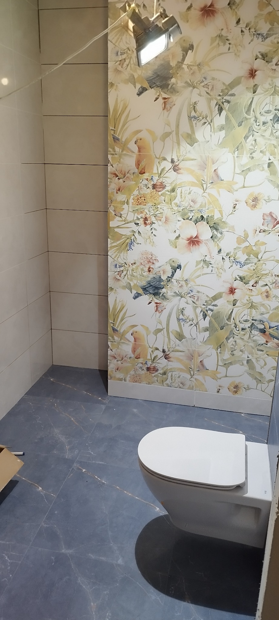 Wnętrze łazienki w trakcie wykańczania: ułożone płytki podłogowe w kolorze granatowym z marmurkowym wzorem, częściowo wyłożone płytki ścienne - białe oraz z motywem kwiatowym i papugami, widoczna...