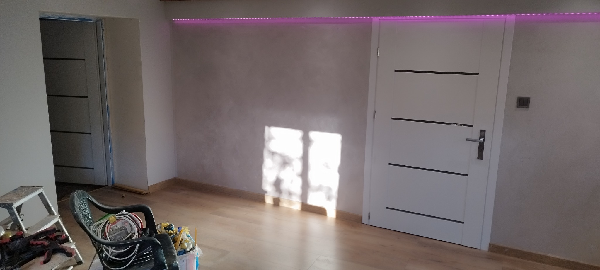 Pokój po odświeżeniu z nową podłogą, pomalowanymi ścianami w odcieniach szarości, białą stolarką drzwiową z poziomymi wstawkami i listwą LED RGB pod sufitem, widoczne narzędzia i drabina malarska.