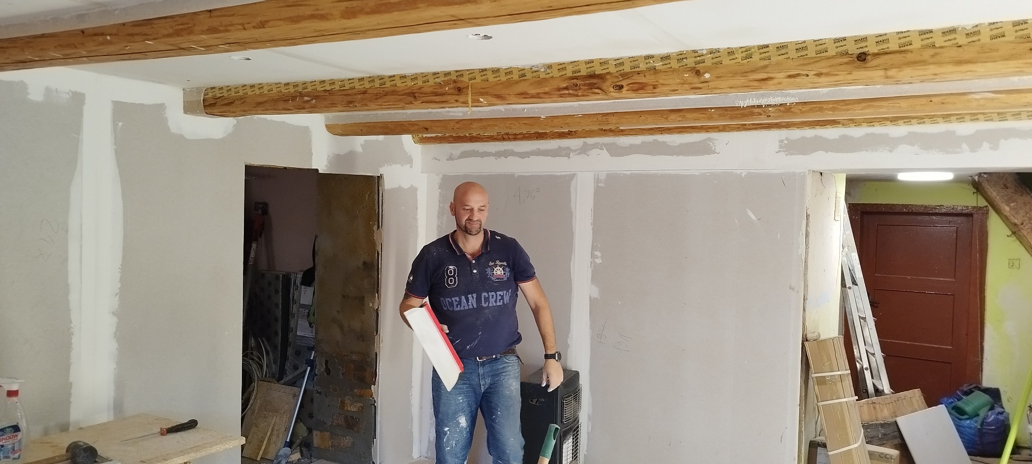 Mężczyzna z pacą szpachlową na tle ściany z płyt kartonowo-gipsowych w trakcie szpachlowania, widoczne drewniane belki stropowe z taśmą zabezpieczającą.