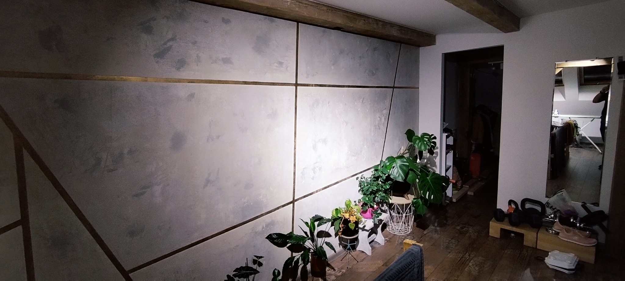 Ściana w salonie z dekoracyjnym wykończeniem w odcieniach szarości i złotymi liniami, drewniany sufit, rośliny doniczkowe i widok na kolejne pomieszczenie w lustrze.