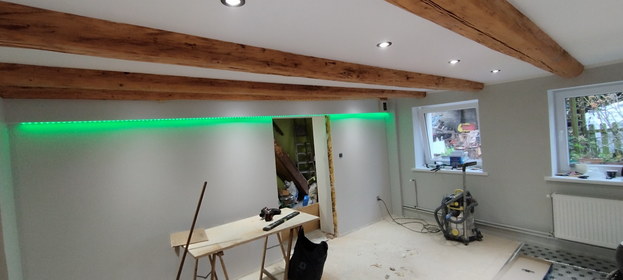 Pokój w trakcie prac wykończeniowych z widocznymi drewnianymi belkami stropowymi, zielonym oświetleniem LED, narzędziami na stole roboczym i odkurzaczem przemysłowym przy oknie.