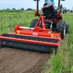 Czerwony traktor z wałem ugniatającym glebę, widok z tyłu, świeżo przygotowany pas ziemi na tle łąki z polnymi kwiatami i zabudowaniami w oddali.