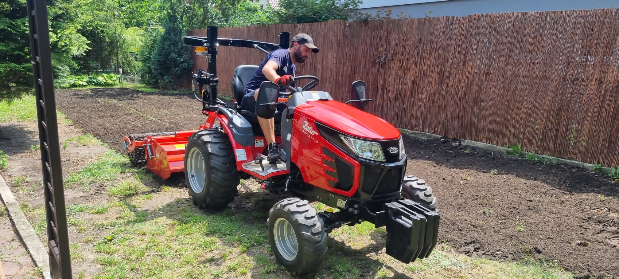 Operator na czerwonym traktorze Zetor Compax HT 25 z glebogryzarką, przygotowujący glebę pod trawnik na tle drewnianego płotu.
