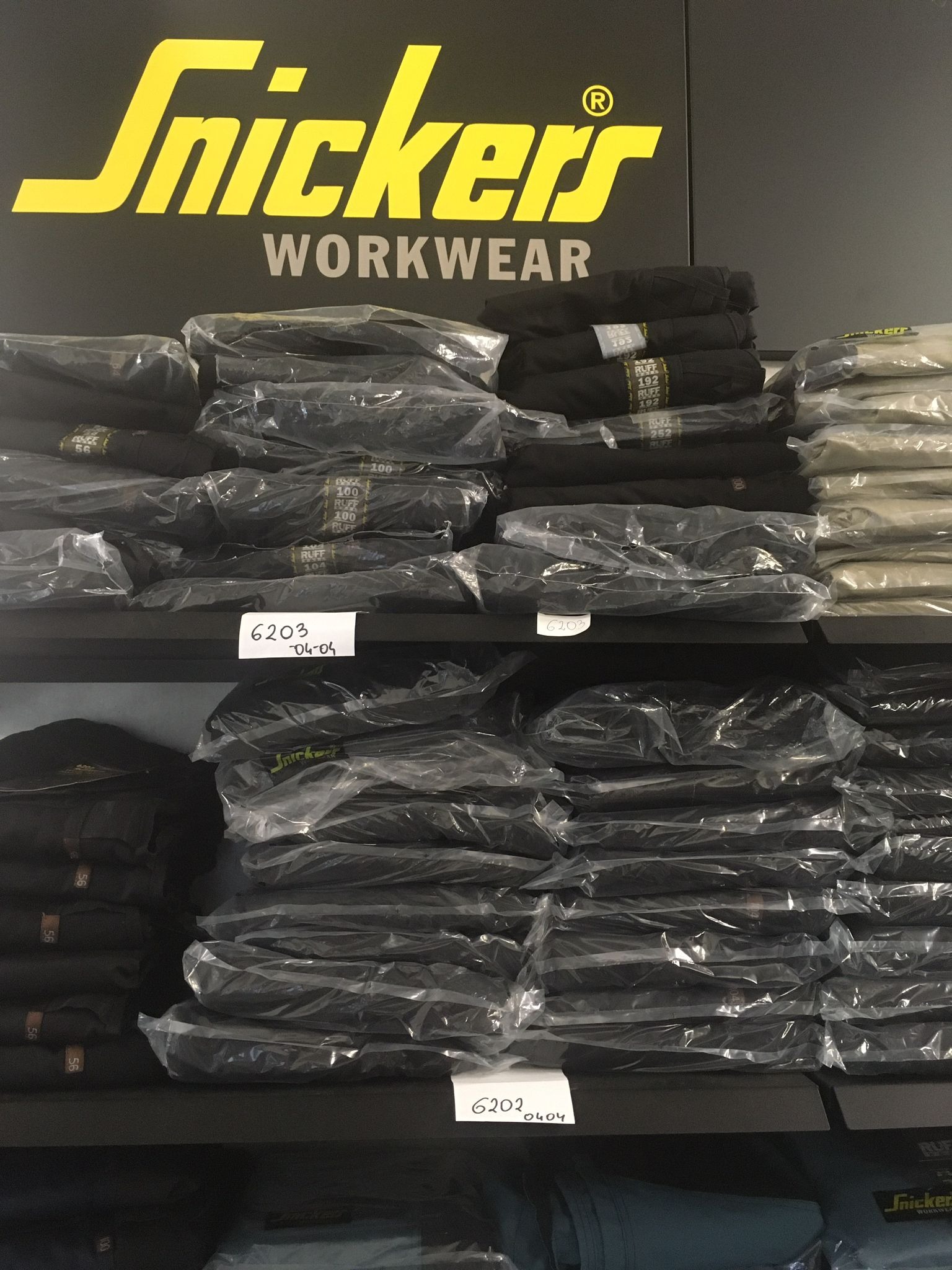 Półki w sklepie z odzieżą roboczą Snickers Workwear, prezentujące stosy zapakowanych ubrań z widocznymi etykietami i oznaczeniami modeli.