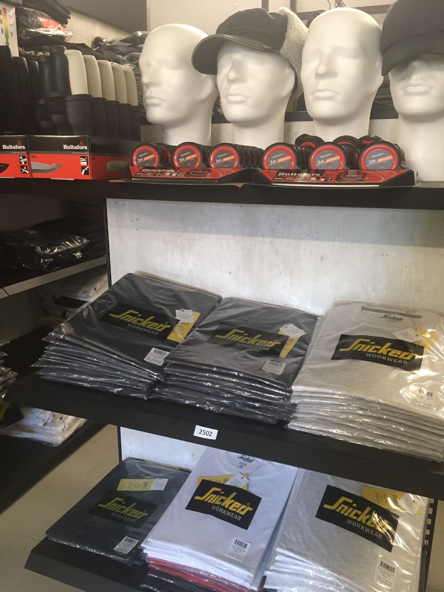 Półki sklepowe z odzieżą roboczą marki Snickers Workwear, manekiny z czapkami i miary zwijane Hultafors na górnej półce.