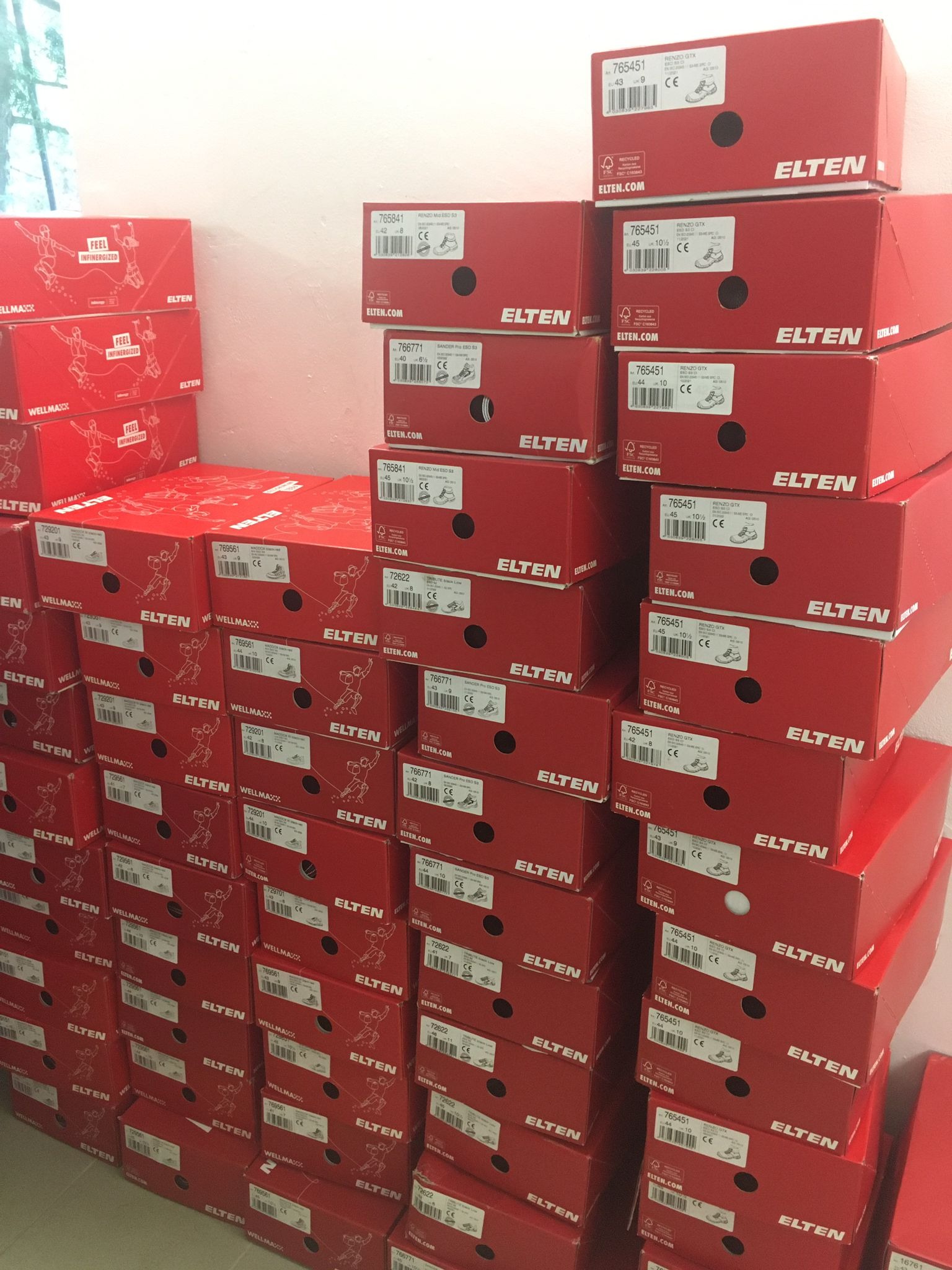 Stos czerwonych pudełek butów roboczych marki Elten, widoczne etykiety z rozmiarami i modelami, ustawione przy białej ścianie.