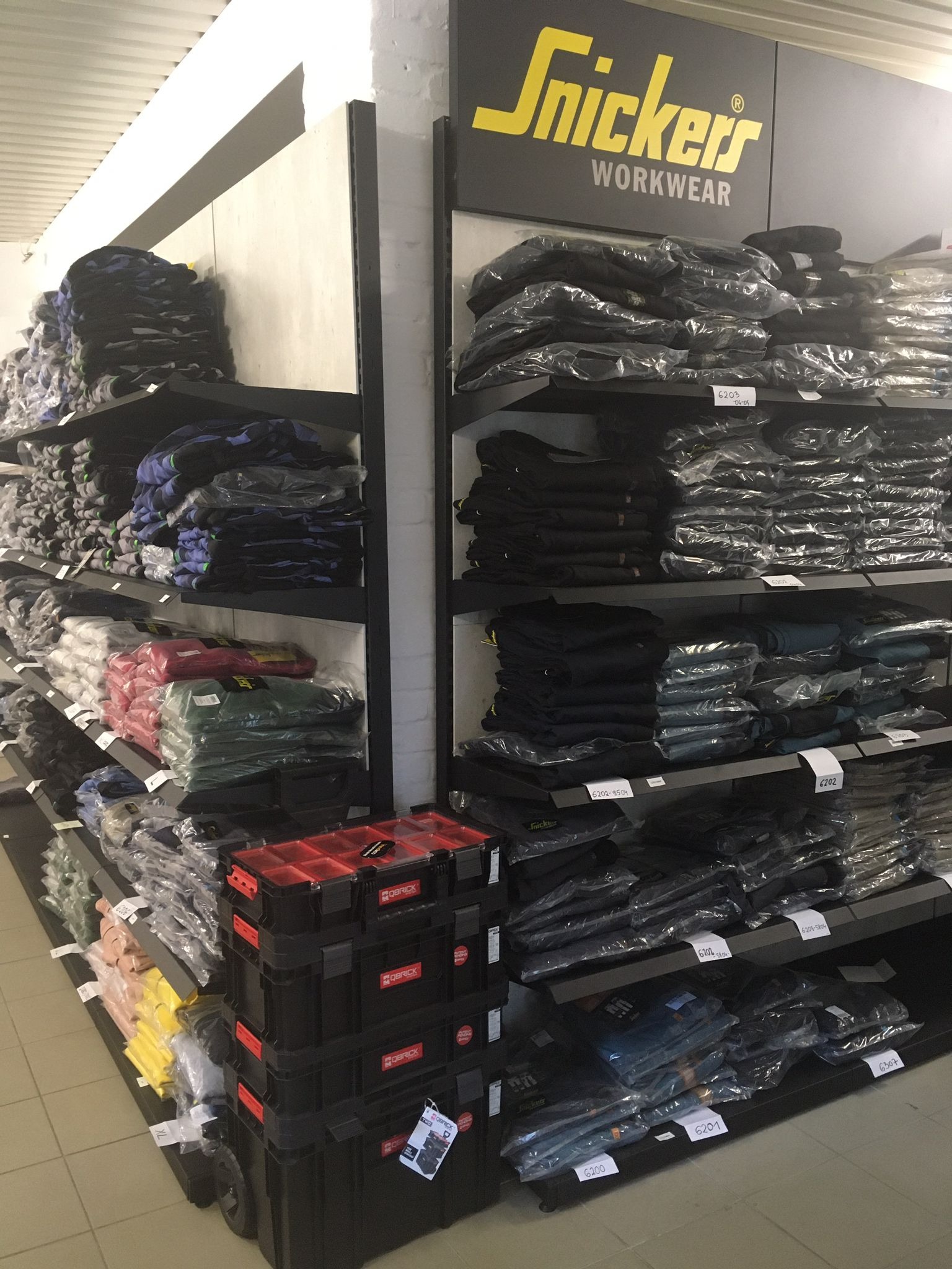 Półki w sklepie z odzieżą roboczą Snickers Workwear, zapakowane ubrania i skrzynki narzędziowe Qbrick na pierwszym planie.