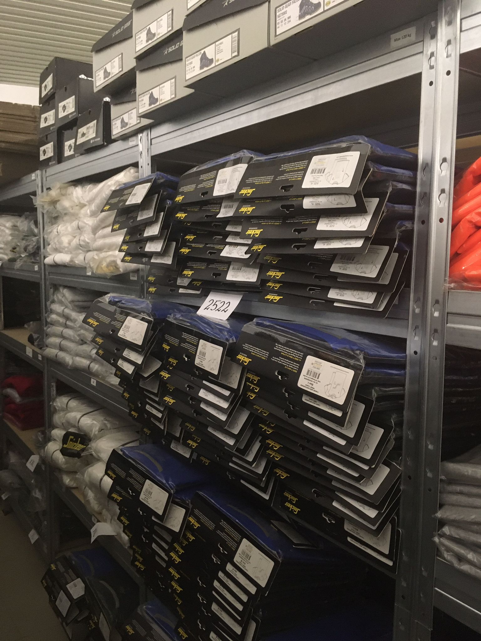 Magazynowe półki z zapasem ubrań roboczych marki Snickers w foliowych opakowaniach, z widocznymi etykietami rozmiarów i numerami katalogowymi, oraz pudełkami butów roboczych na górnej półce.