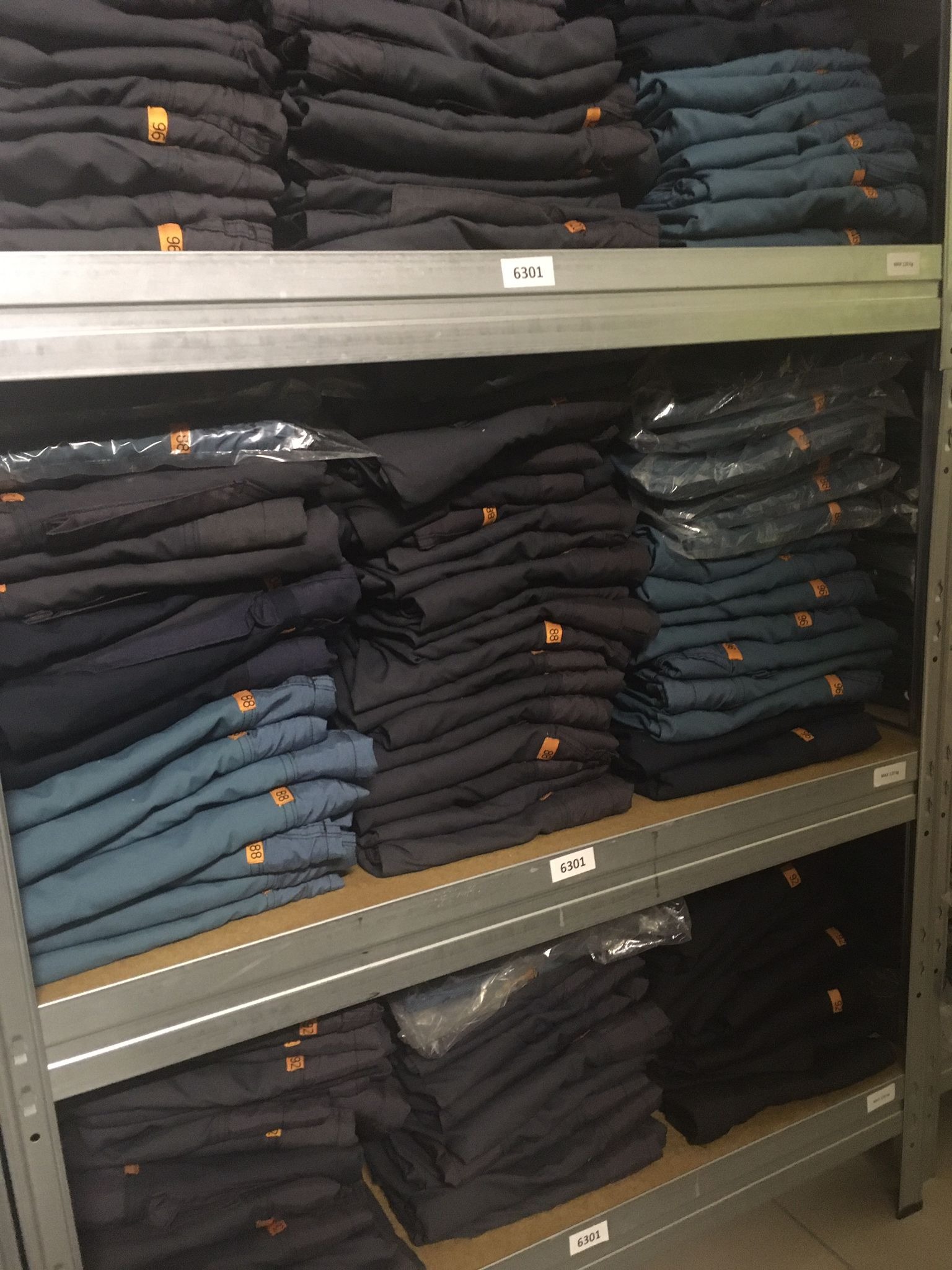 Metalowy regał magazynowy wypełniony złożoną odzieżą roboczą w odcieniach granatu i błękitu, każda sztuka oznaczona pomarańczową etykietą z numerem, część ubrań zapakowana w folię.