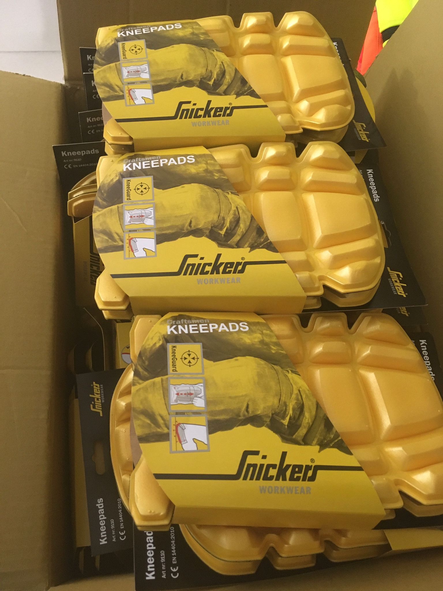 Stos żółtych nakolanników Snickers Workwear w kartonowym pudełku, widoczne opakowania z instrukcją montażu.