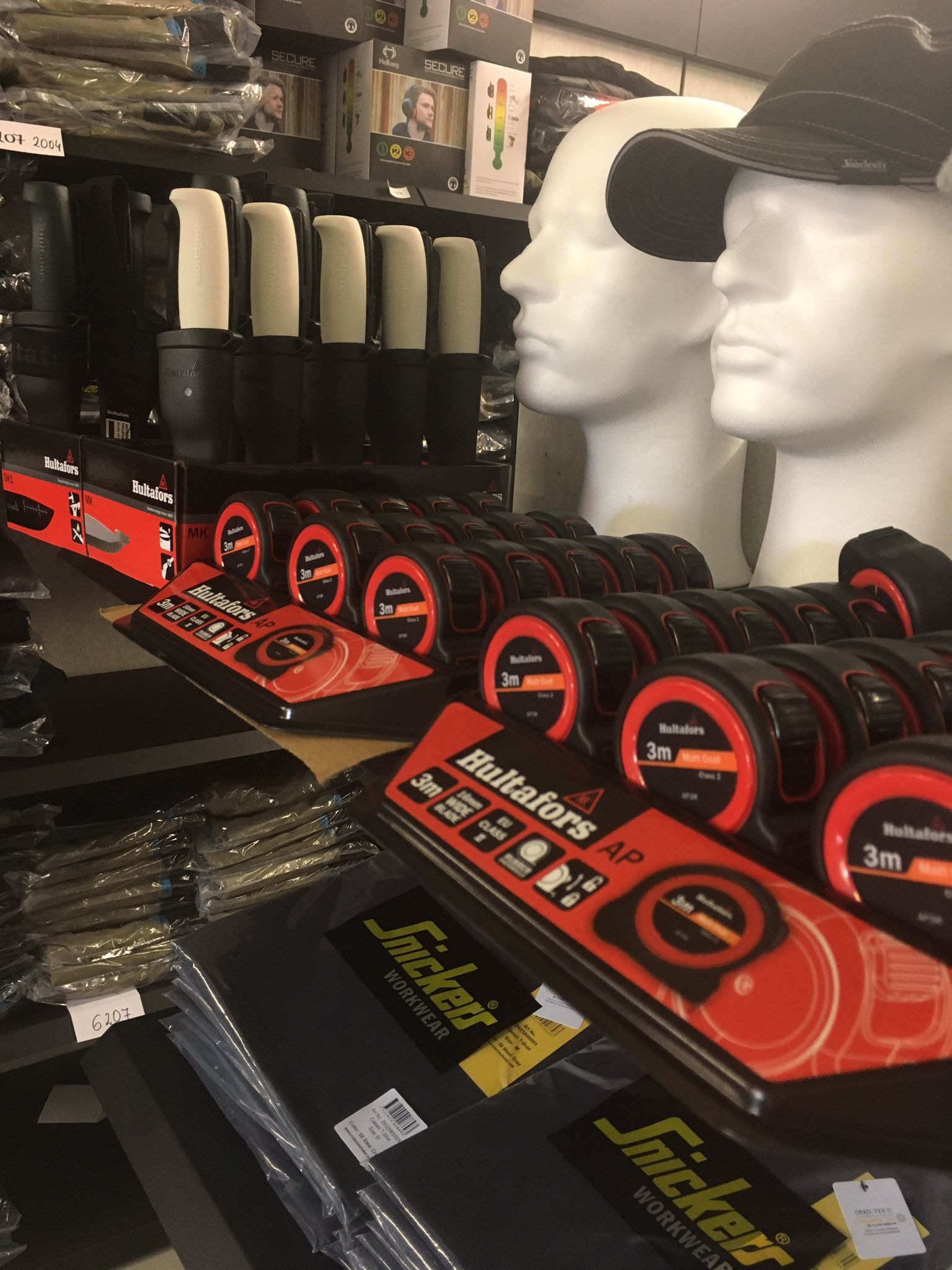 Ekspozycja w sklepie z odzieżą roboczą: Manekiny w czapkach, miary zwijane Hultafors, noże Hultafors i zapakowana odzież robocza Snickers Workwear na półkach.