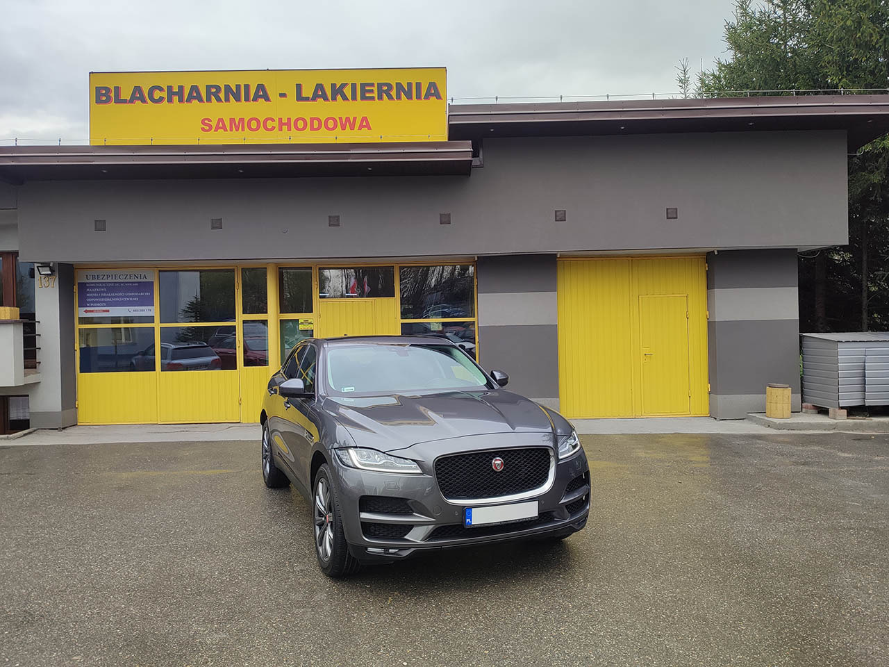Szary Jaguar F-Pace zaparkowany przed budynkiem warsztatu z żółtymi drzwiami i szyldem informującym o usługach naprawy karoserii samochodowej.