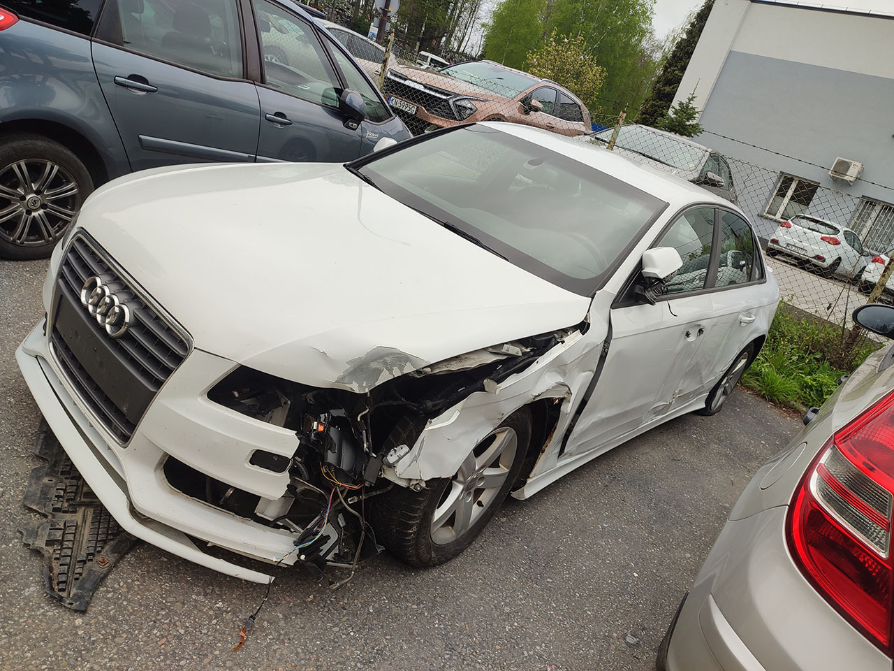 Białe Audi A4 z poważnymi uszkodzeniami przodu po kolizji, widoczne zniszczone elementy karoserii i wystające przewody, na parkingu wśród innych pojazdów.