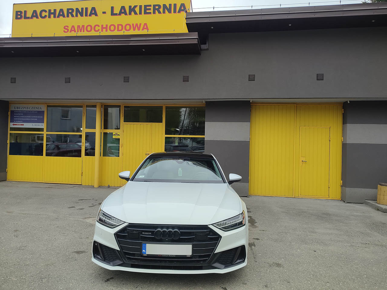 Białe Audi A7 zaparkowane przed budynkiem warsztatu z żółtymi drzwiami i napisem 'Blacharnia - Lakiernia Samochodowa' na żółtym szyldzie.