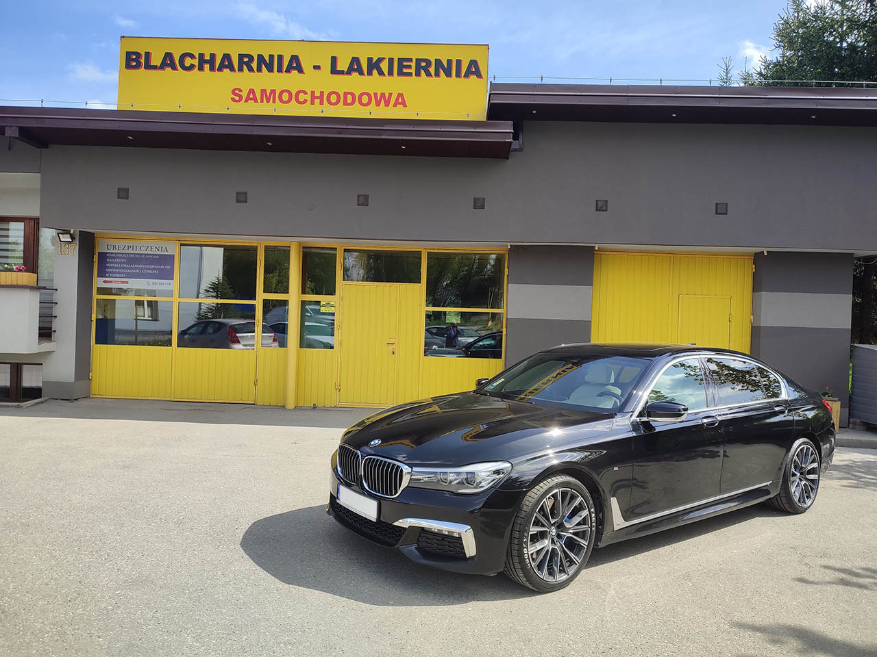 Czarny samochód BMW serii 7 zaparkowany przed budynkiem blacharni-lakierni z żółtymi bramami garażowymi i szyldem reklamowym.