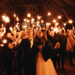 Uśmiechnięta para młoda otoczona gośćmi z zimnymi ogniami pod drewnianą konstrukcją dachu. Radosna atmosfera świętowania.