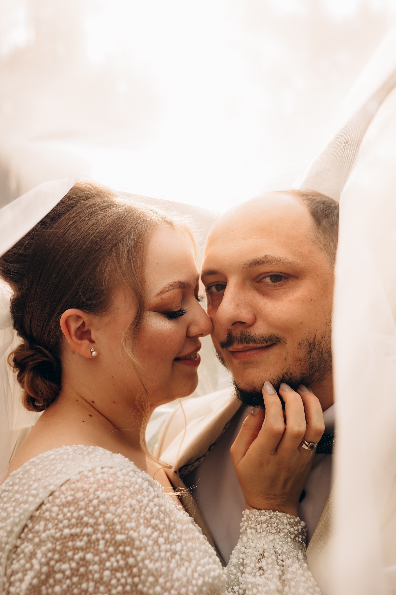 Panna młoda i pan młody w zbliżeniu, otuleni welonem, ona z delikatnym makijażem i perłowymi kolczykami, on z ręką na jej policzku, oboje uśmiechnięci.