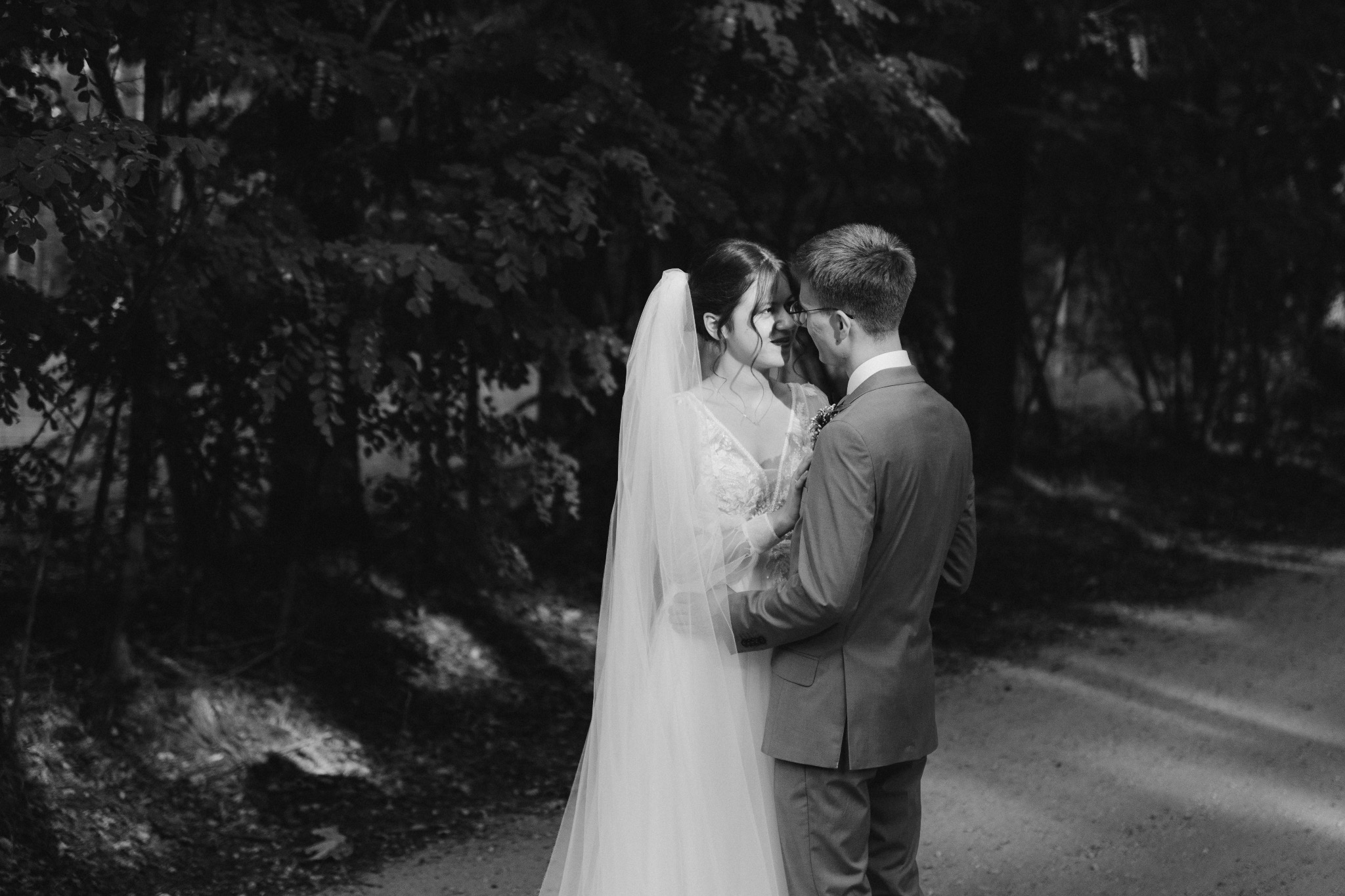 Czarno-białe zdjęcie pary młodej w plenerze: panna młoda w koronkowej sukni i długim welonie, pan młody w garniturze, oboje patrzą na siebie z uśmiechem na tle drzew i ścieżki.