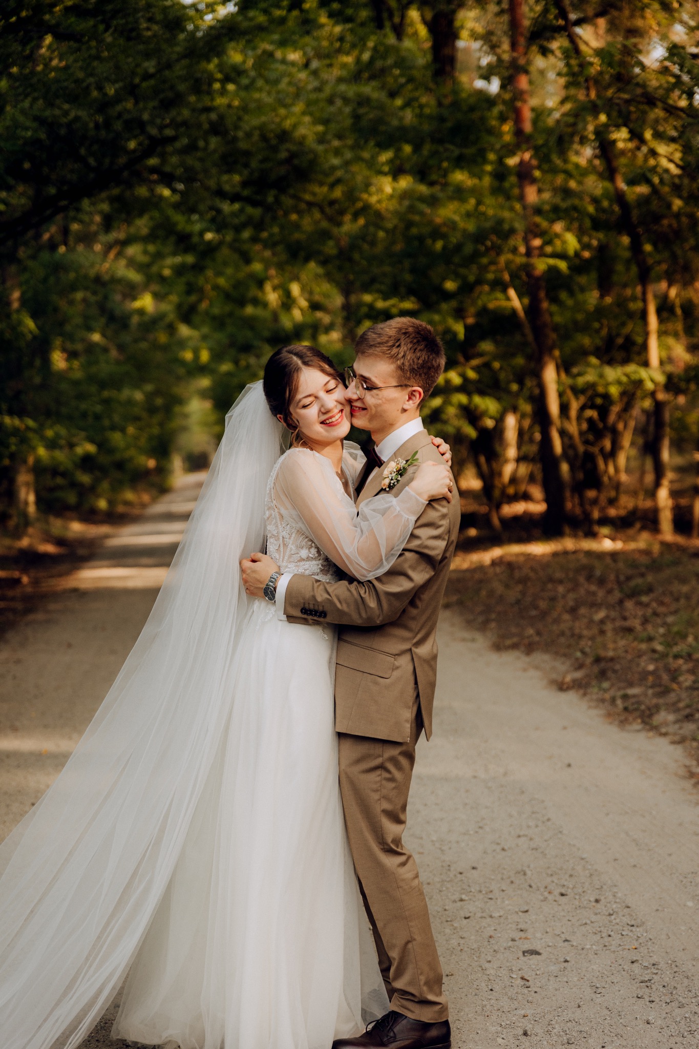 Uśmiechnięta panna młoda w koronkowej sukni z długim welonem, obejmowana przez pana młodego w brązowym garniturze na leśnej drodze.