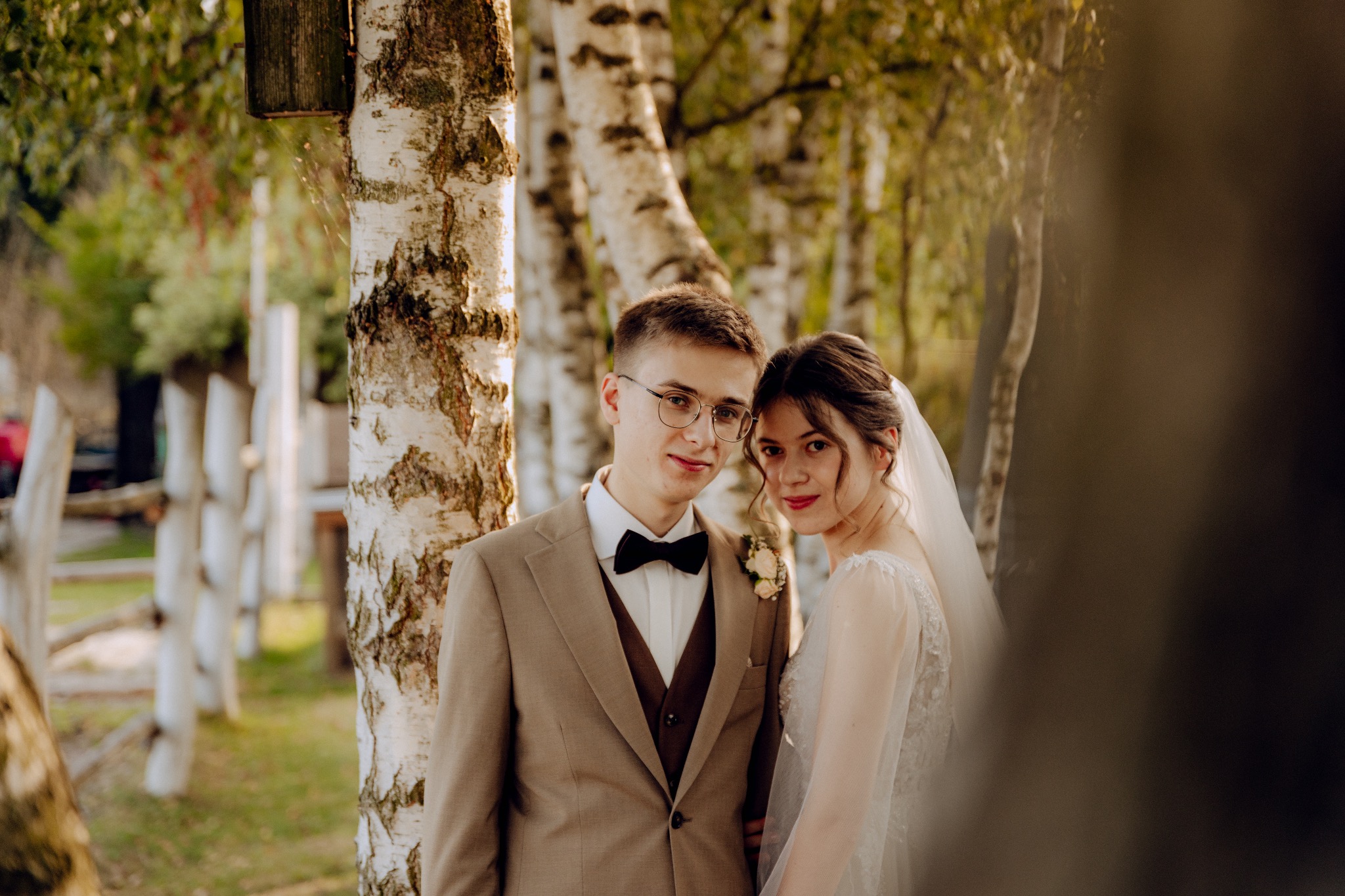 Para młoda w lesie brzozowym: pan młody w beżowym garniturze i czarnej muszce, panna młoda w białej sukni z welonem, oboje uśmiechnięci, delikatne światło.