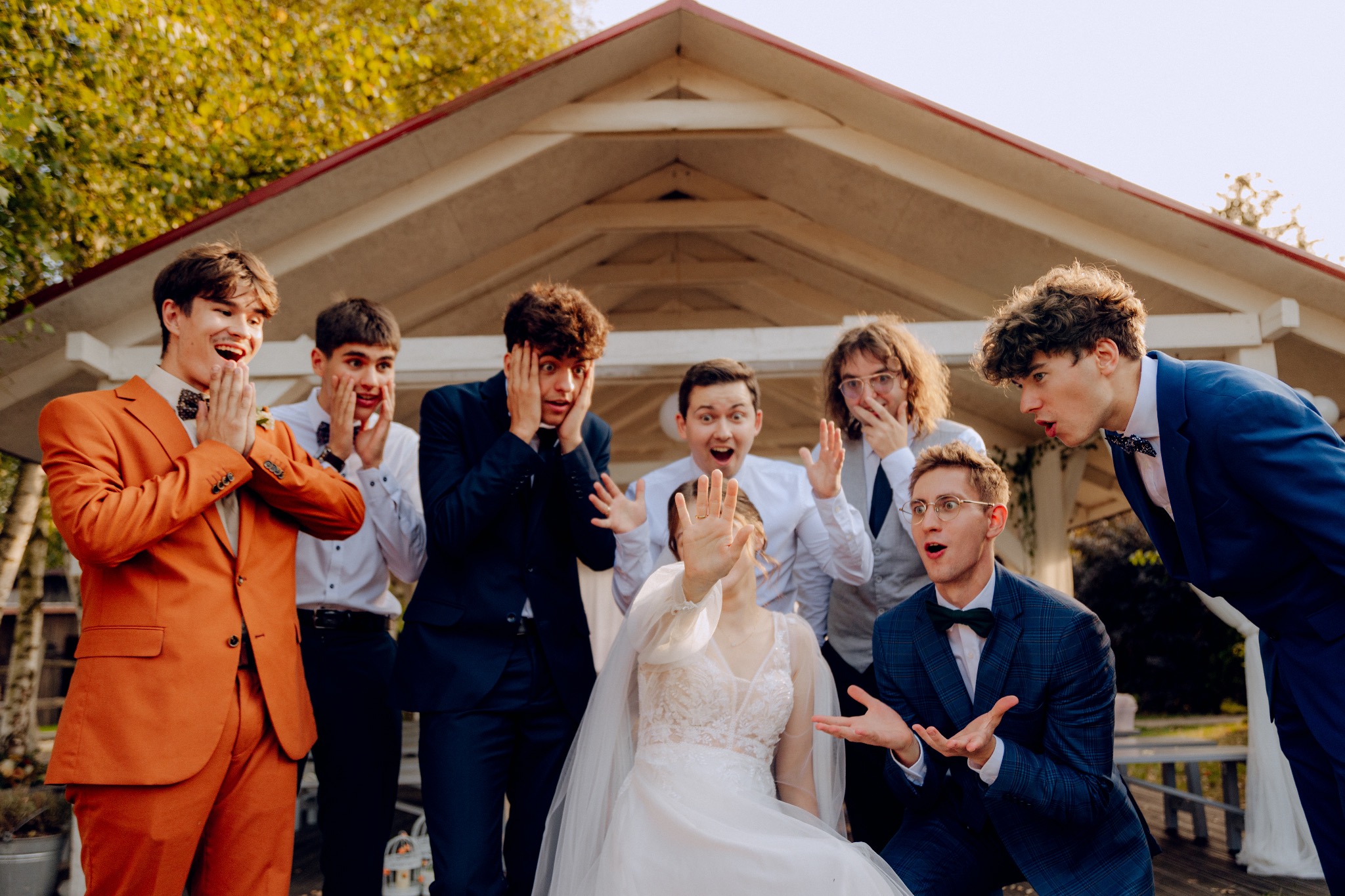 Panna młoda zasłaniająca twarz dłonią z pierścionkiem, otoczona zaskoczonymi, elegancko ubranymi mężczyznami na tle białej altany.