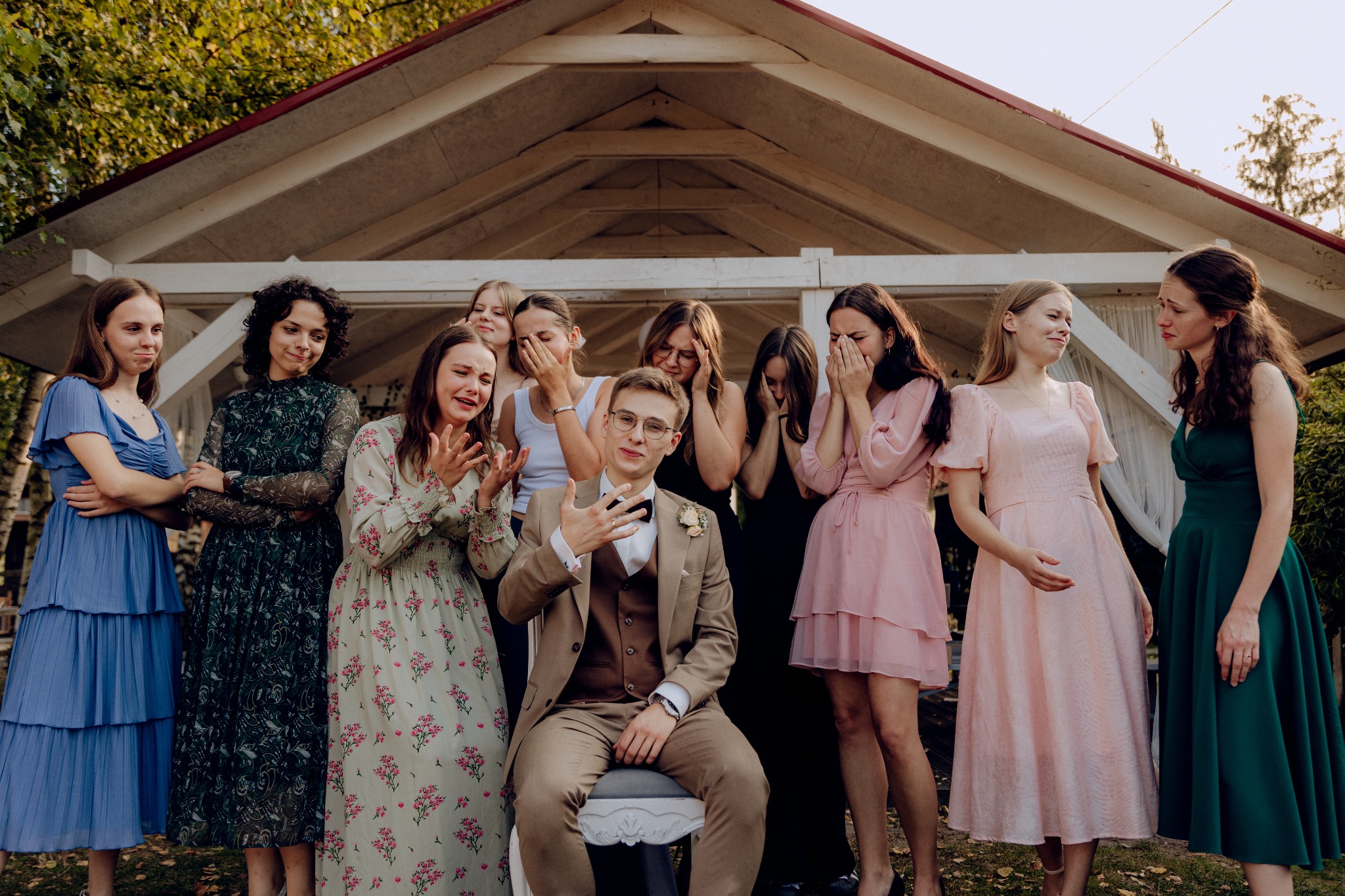 Pan młody siedzi na krześle otoczony przez wzruszone druhny w sukienkach w odcieniach zieleni, błękitu i różu, na tle białej altany.