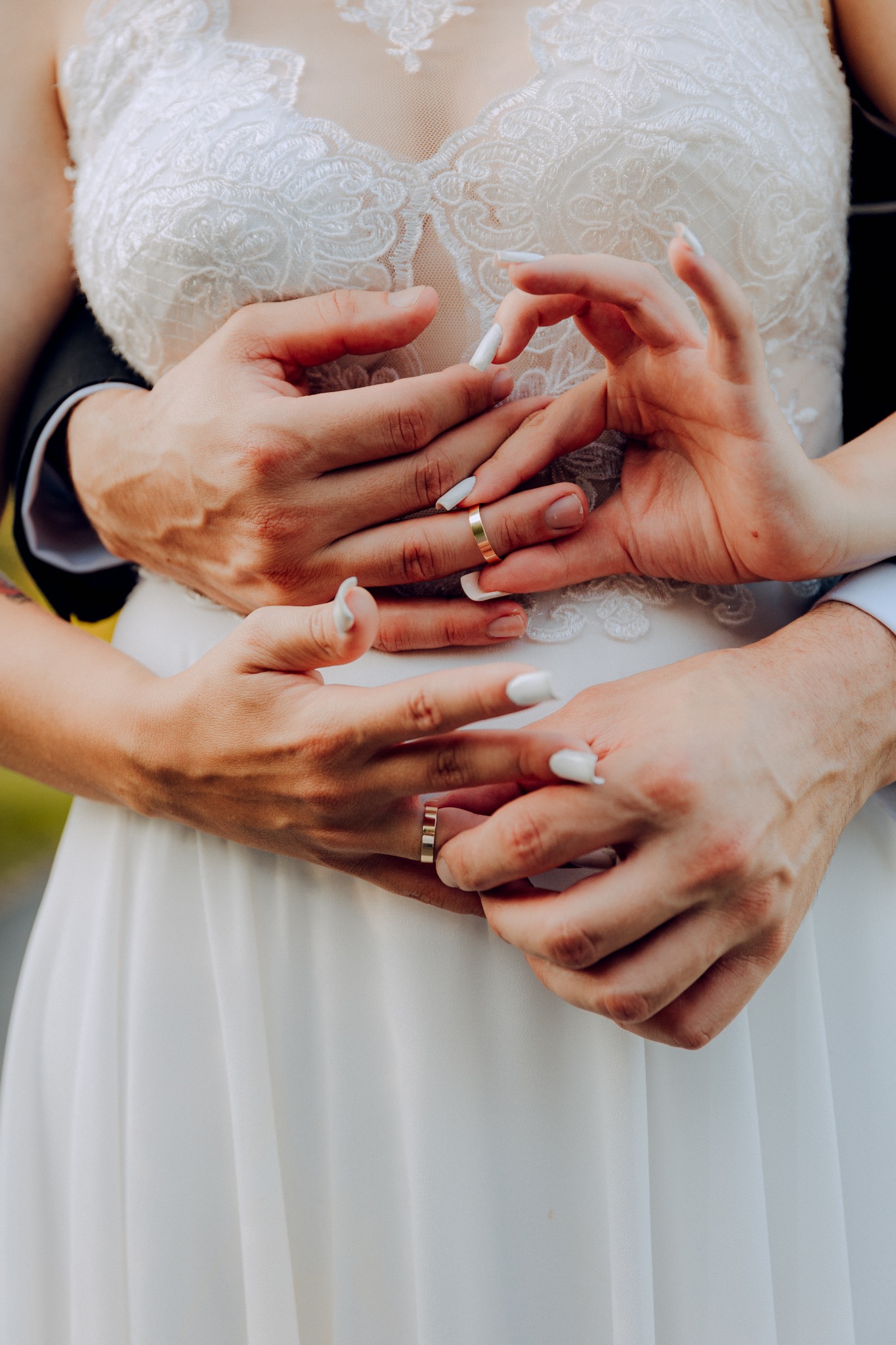 Dłonie nowożeńców z złotymi obrączkami na tle białej, koronkowej sukni ślubnej.