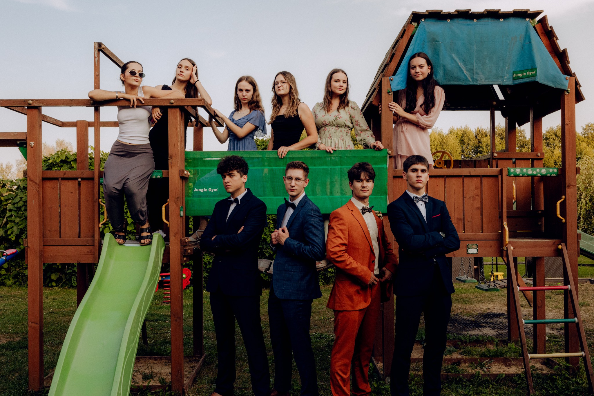 Grupa nastolatków w strojach galowych i sukienkach pozuje na tle placu zabaw Jungle Gym z zieloną zjeżdżalnią i drewnianą konstrukcją.