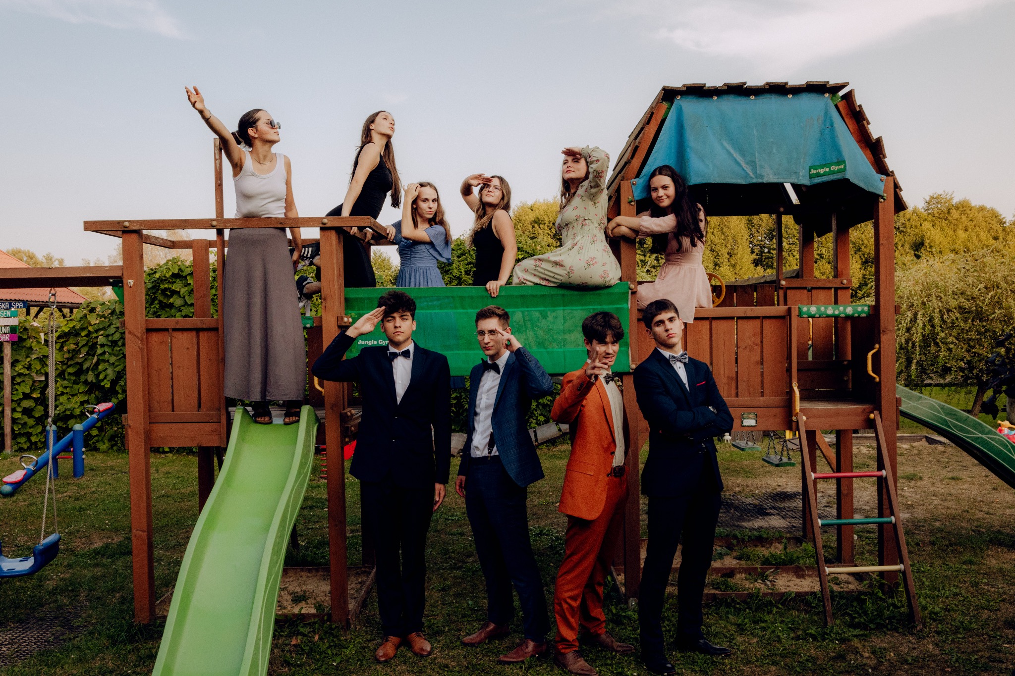 Grupa młodzieży w strojach galowych pozuje na tle drewnianego placu zabaw w ogrodzie podczas sesji fotograficznej.