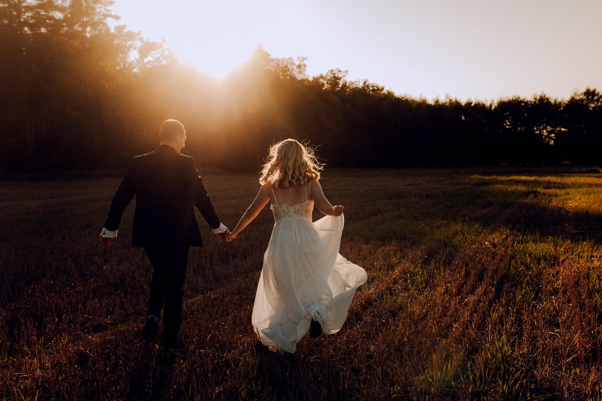 Para młoda, trzymając się za ręce, biegnie przez pole w promieniach zachodzącego słońca, sukienka panny młodej powiewa na wietrze.