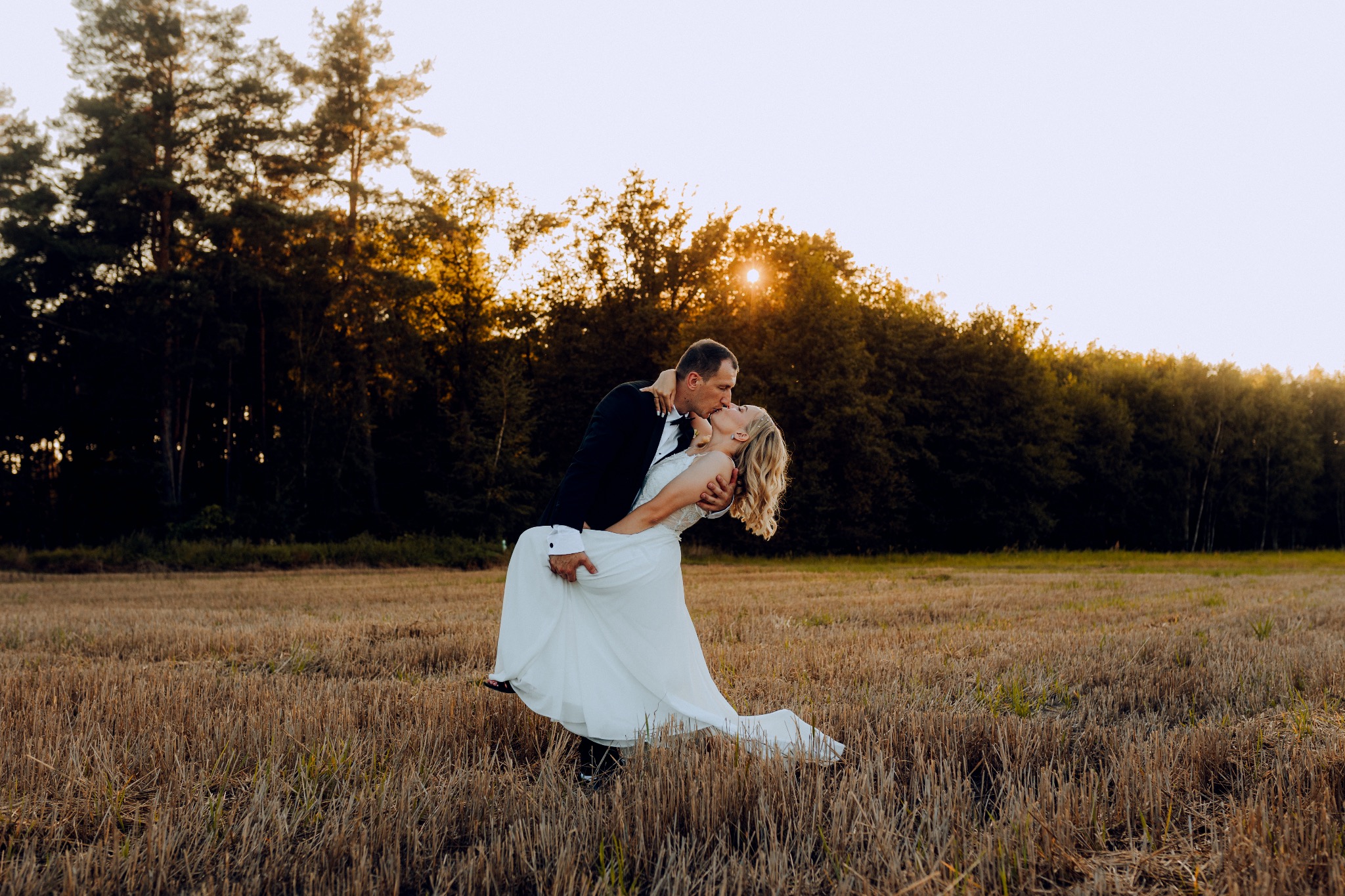 Panna młoda w białej sukni ślubnej, unoszona przez pana młodego w smokingu, podczas pocałunku na polu ścierniska w blasku zachodzącego słońca.