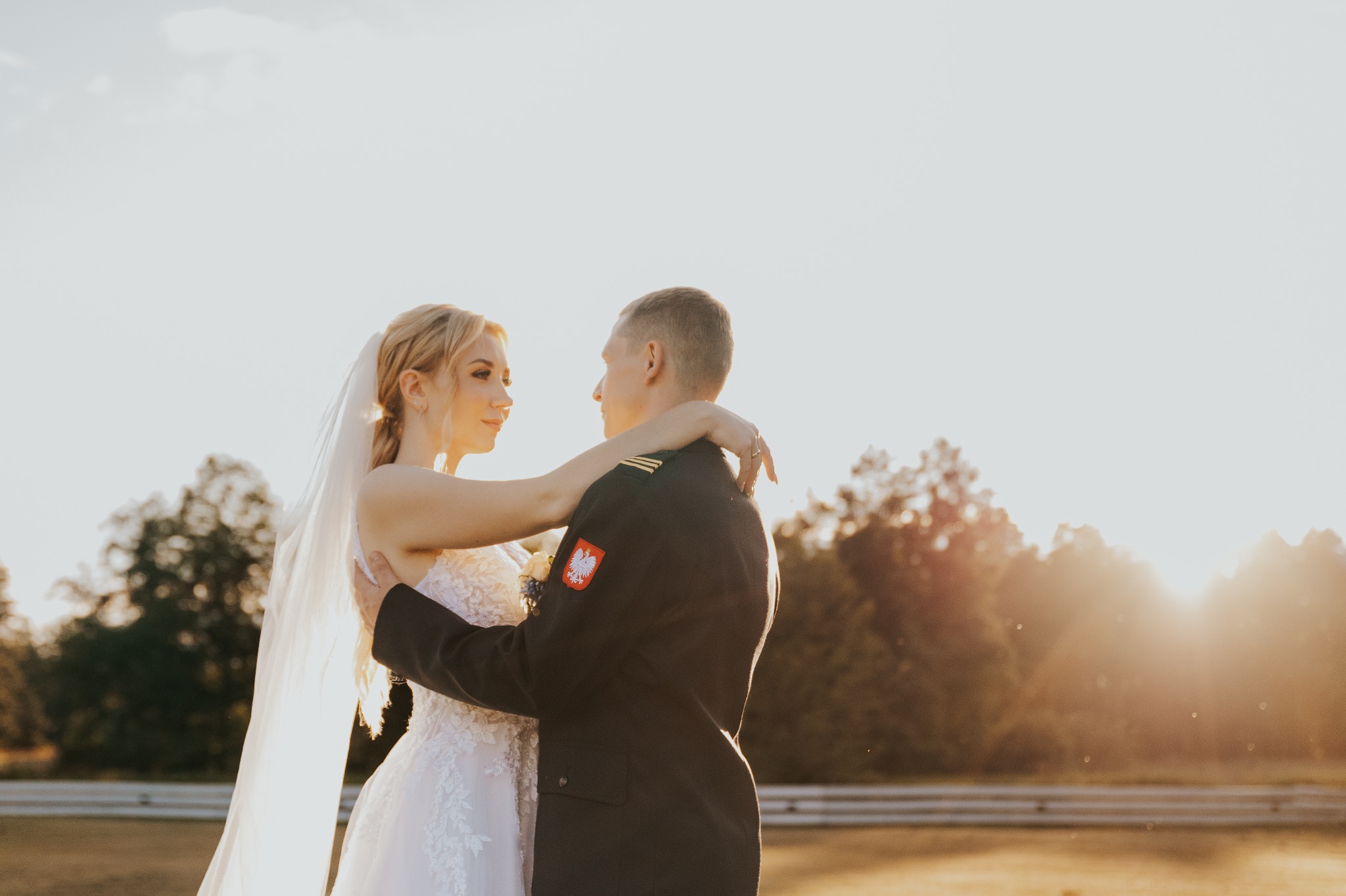 Panna młoda w koronkowej sukni i welonie obejmuje oficera w mundurze Wojska Polskiego w promieniach zachodzącego słońca.