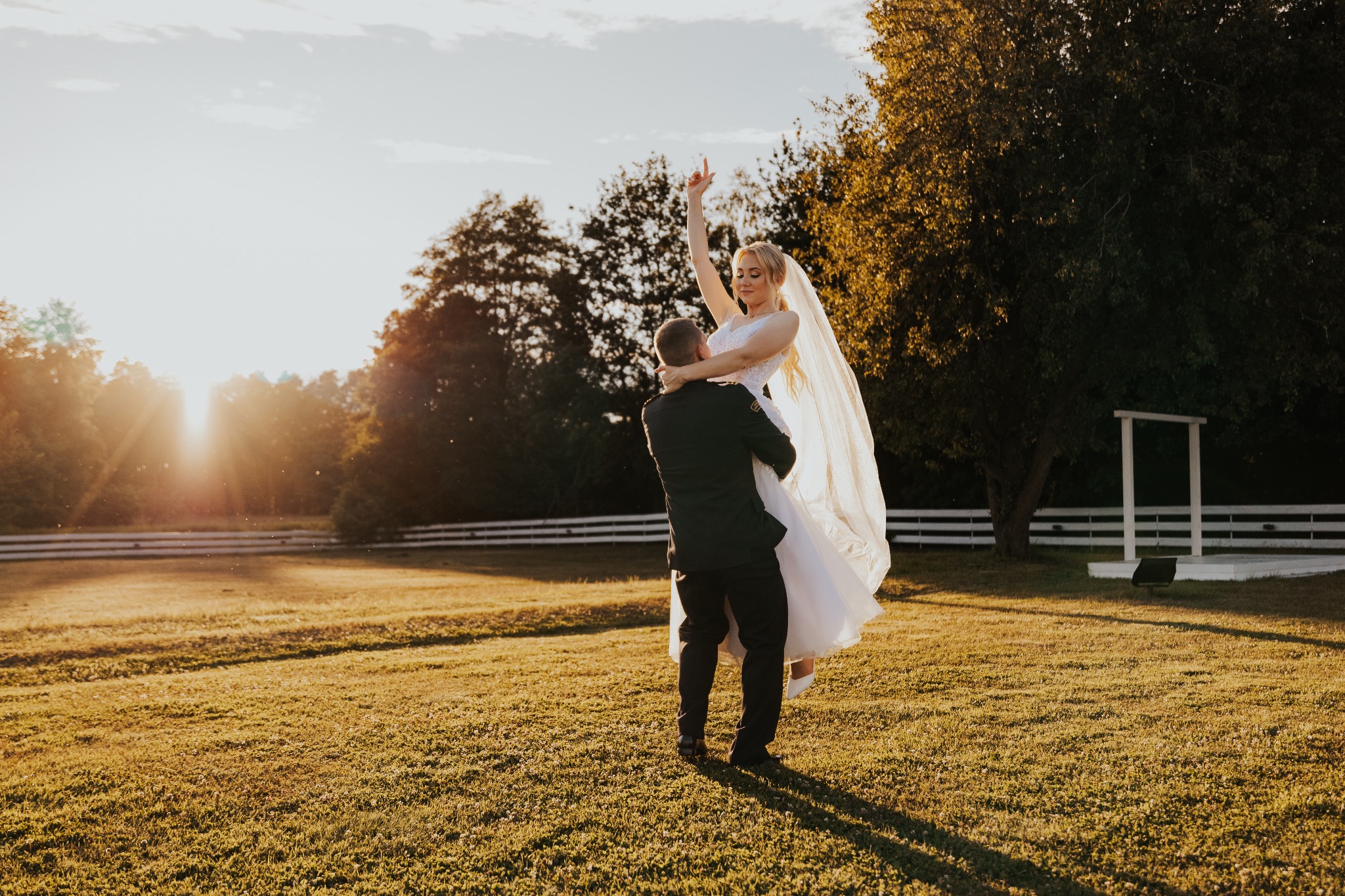 Pan młody podnosi pannę młodą w białej sukni i welonie na tle zachodzącego słońca i zielonego trawnika.